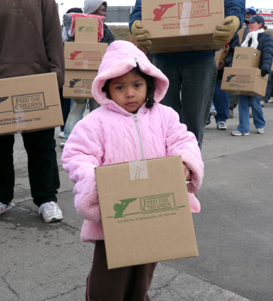 Una niña pequeña con un abrigo rosa sostiene una caja en un evento al aire libre