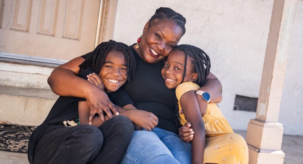 Una madre y sus dos hijos se abrazan sentados en el porche de una casa