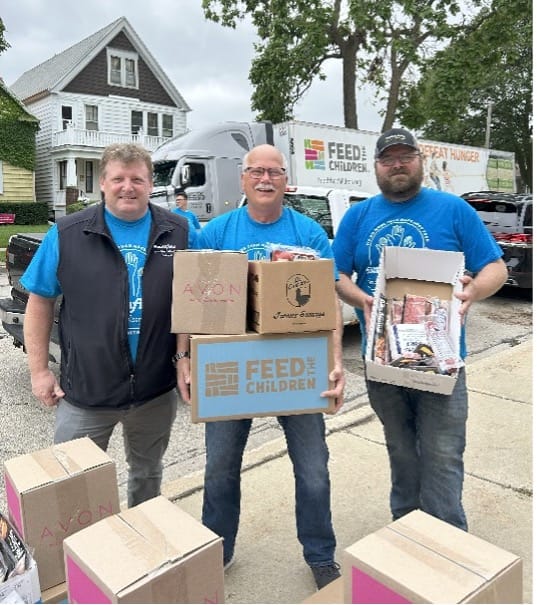 Three men holding Feed the Children boxes at an outdoor event.