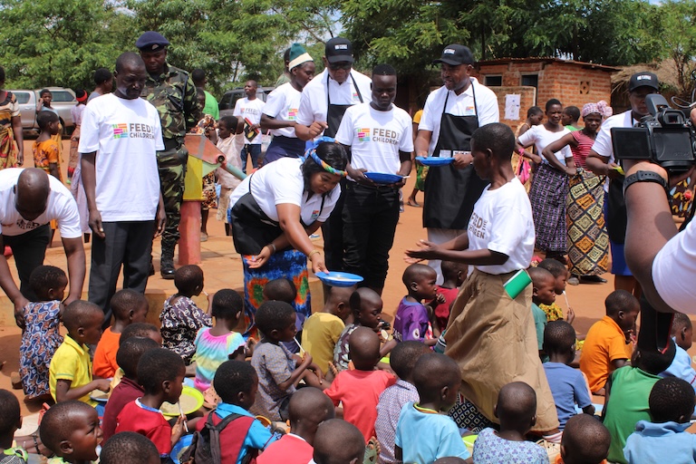 An outdoor event with a crowd of adults handing out food to children.