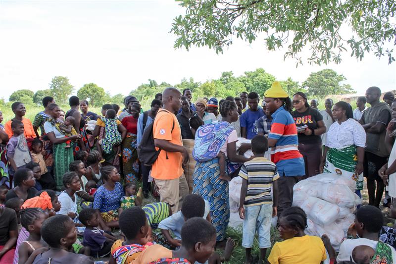 A large group of community members gathered beneath the shade of tall trees.