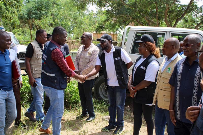 A group of aid workers and local officials stand together outdoors near parked vehicles.